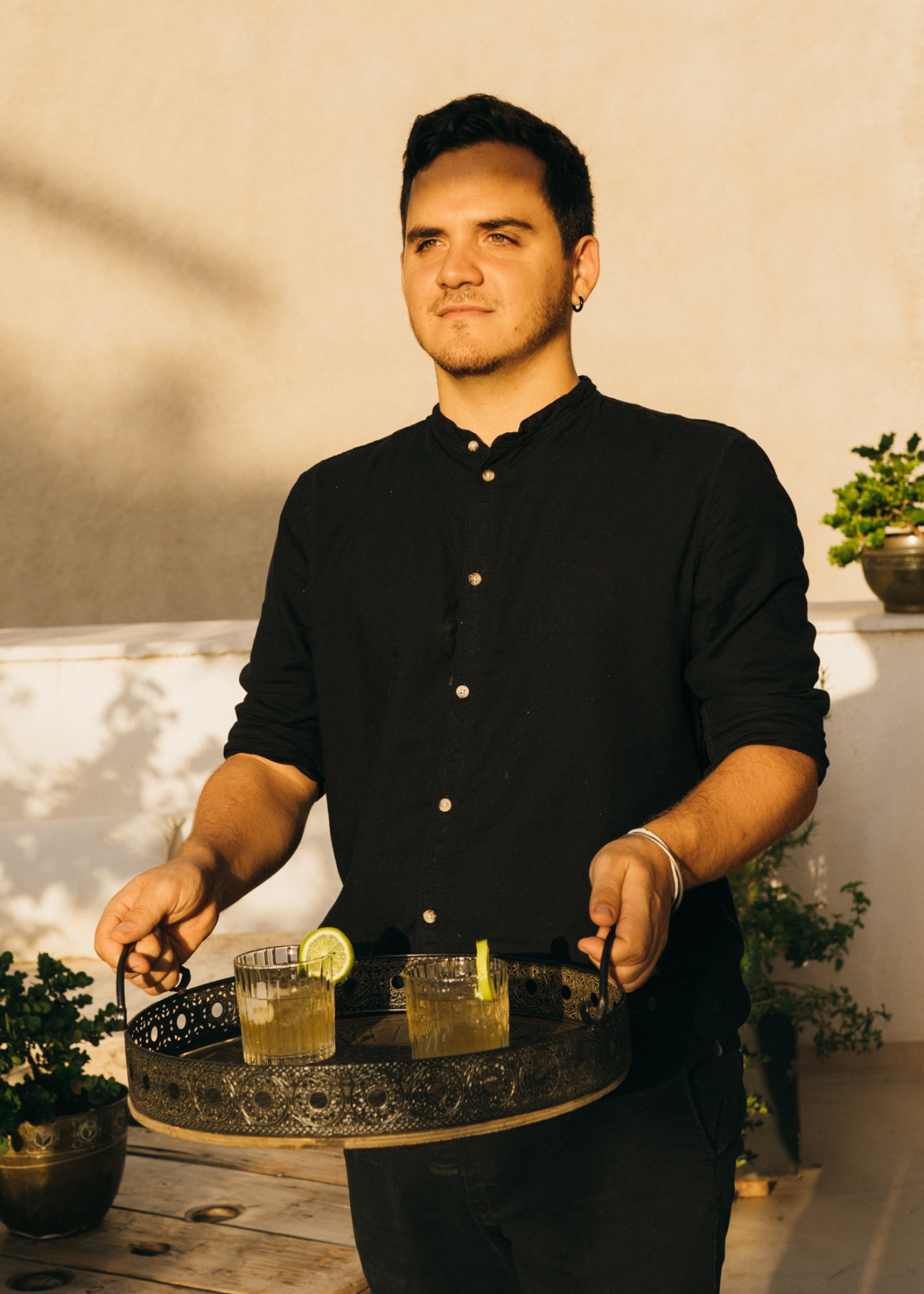 Man in black shirt serving cocktails on decorative metal tray in elegant interior with plants