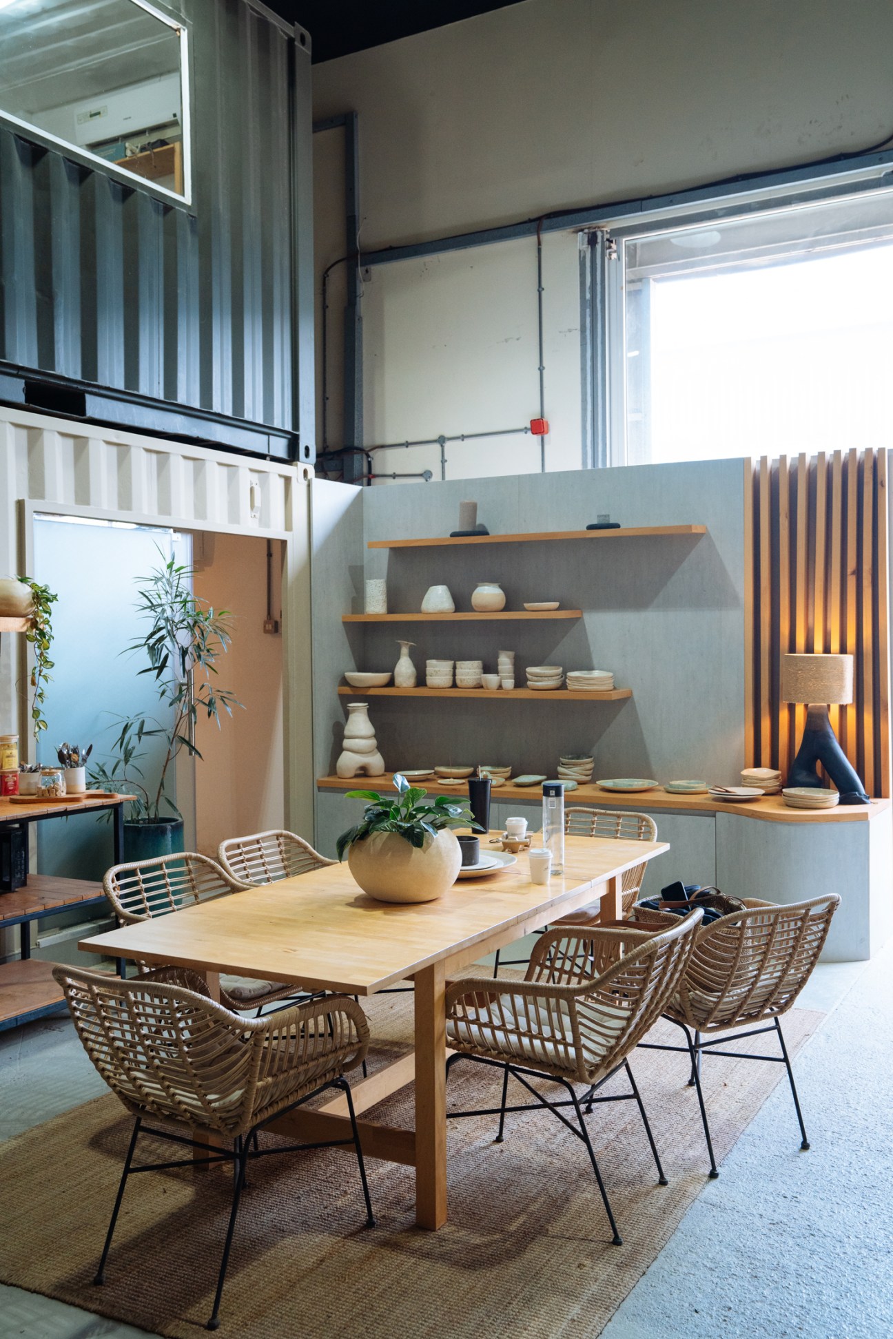 A modern studio space with a wooden dining table, woven chairs, and built-in shelving displaying ceramic vessels and bowls.