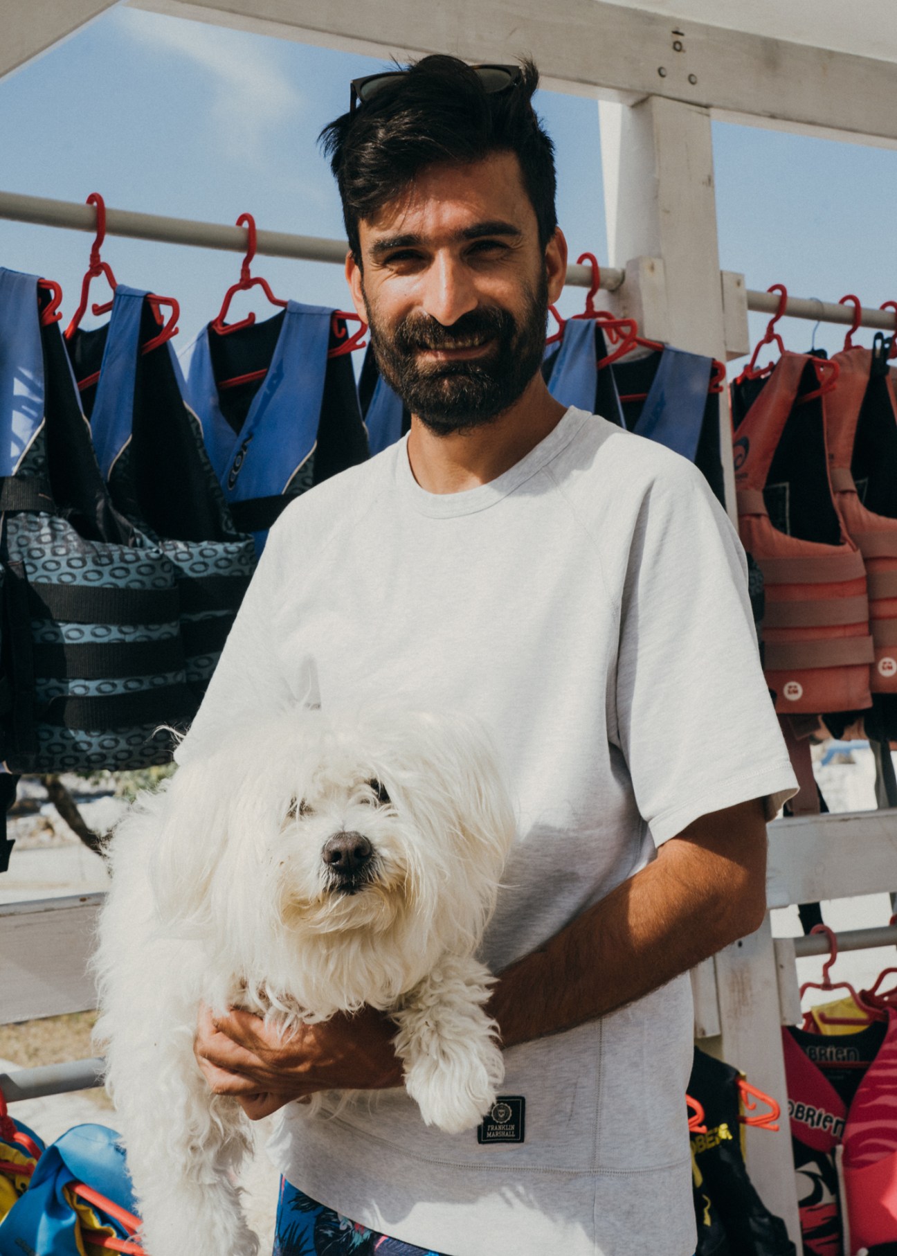 Vassilis Lagopoulos holds his white dog Rocky while standing in front of water sports equipment on a sunny beach day.