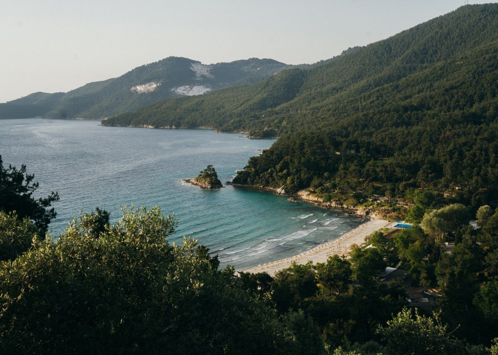 Aerial view of Makryammos beach on Thassos with forested mountains, turquoise sea, and sandy shoreline.