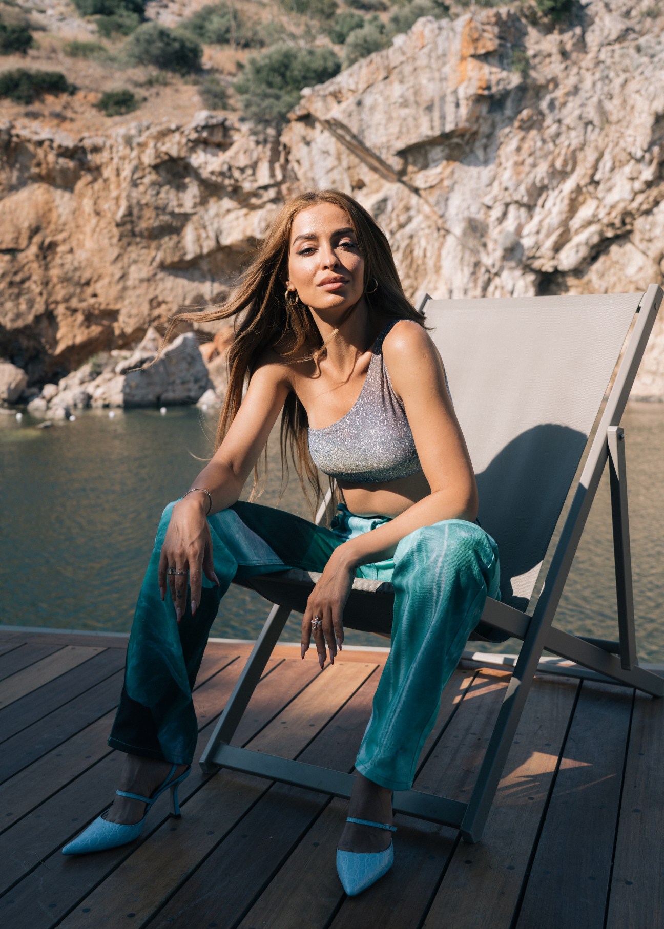 Woman sitting on a white deck chair by a rocky coastline, wearing a grey crop top and teal pants.