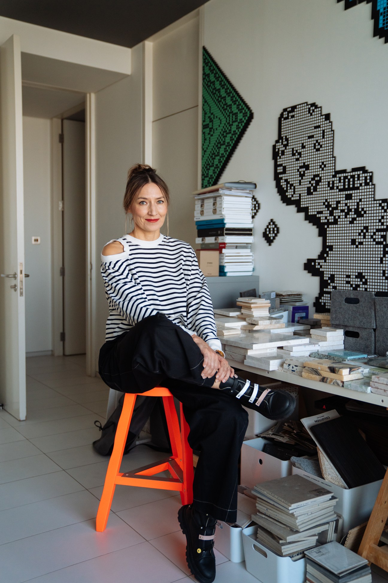 Designer Agata Kurzela sits on an orange stool in her minimalist studio surrounded by books and pixel art on the wall.