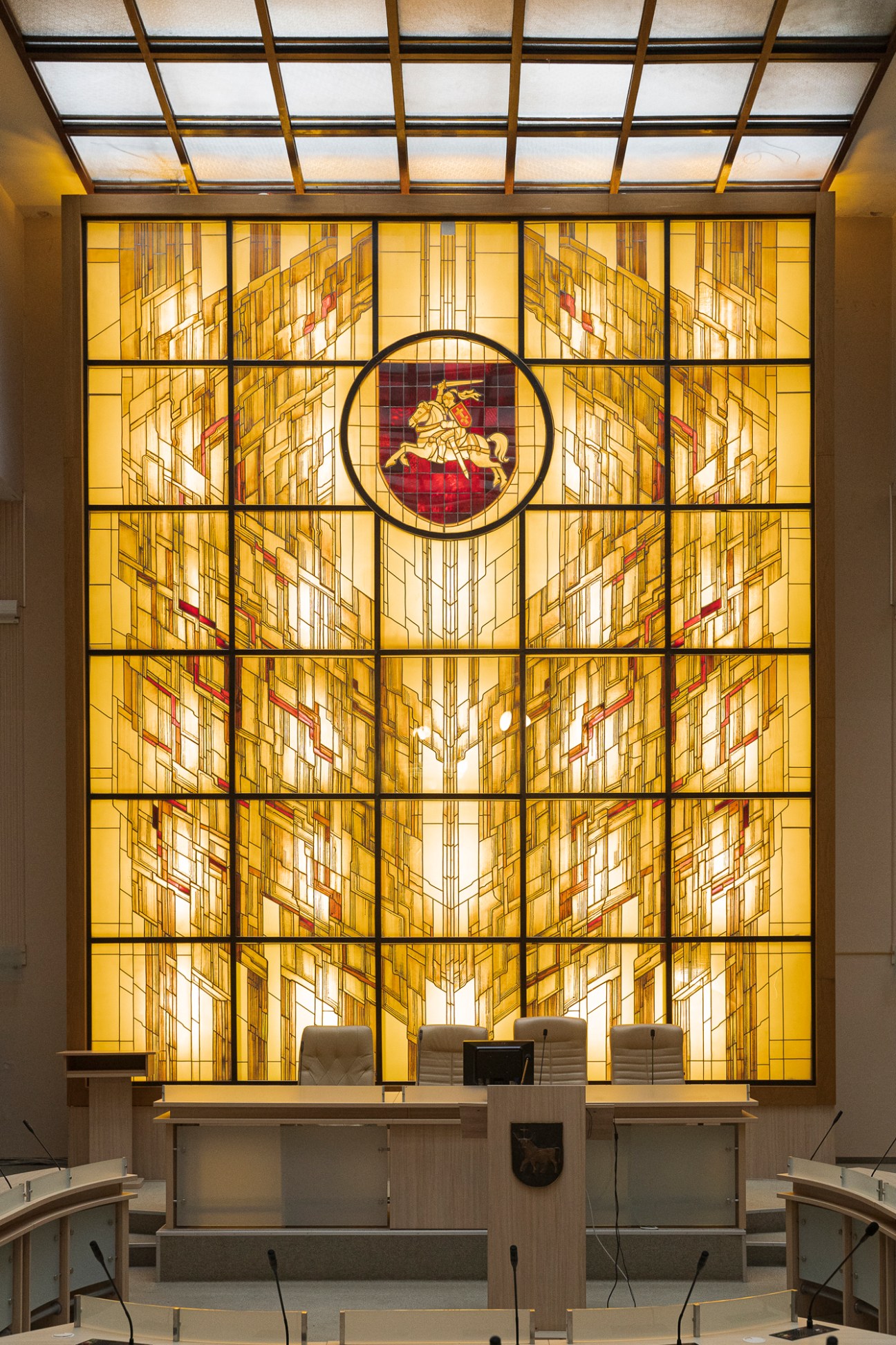 Ornate stained glass window with geometric patterns and central coat of arms in municipal building interior