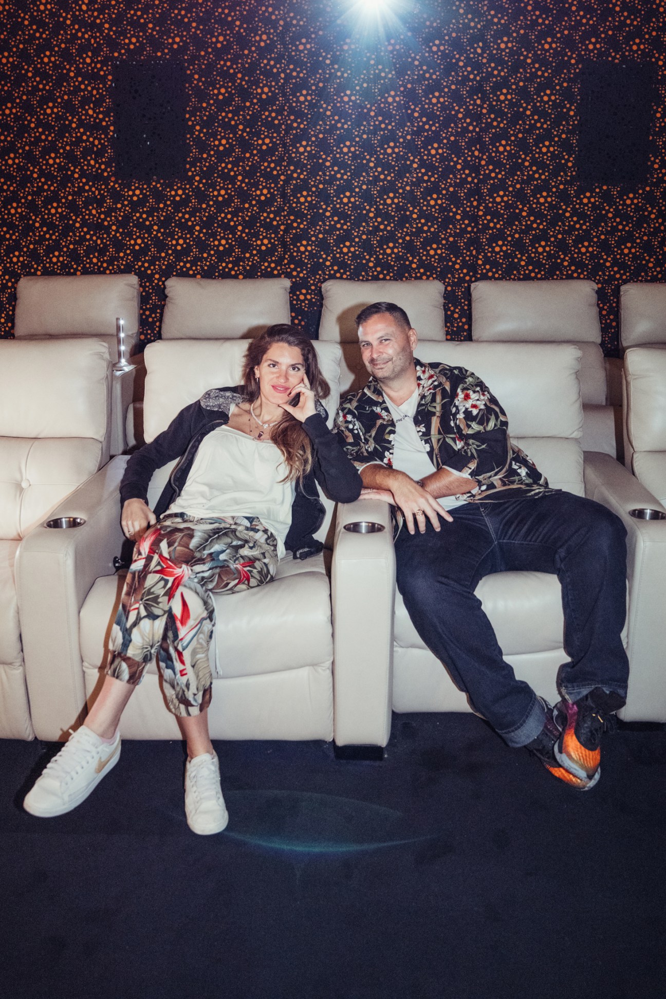 Hassiba and Kenton Freiha-Oxley sit together on a cream-colored theater seat against an illuminated orange and dark patterned backdrop.