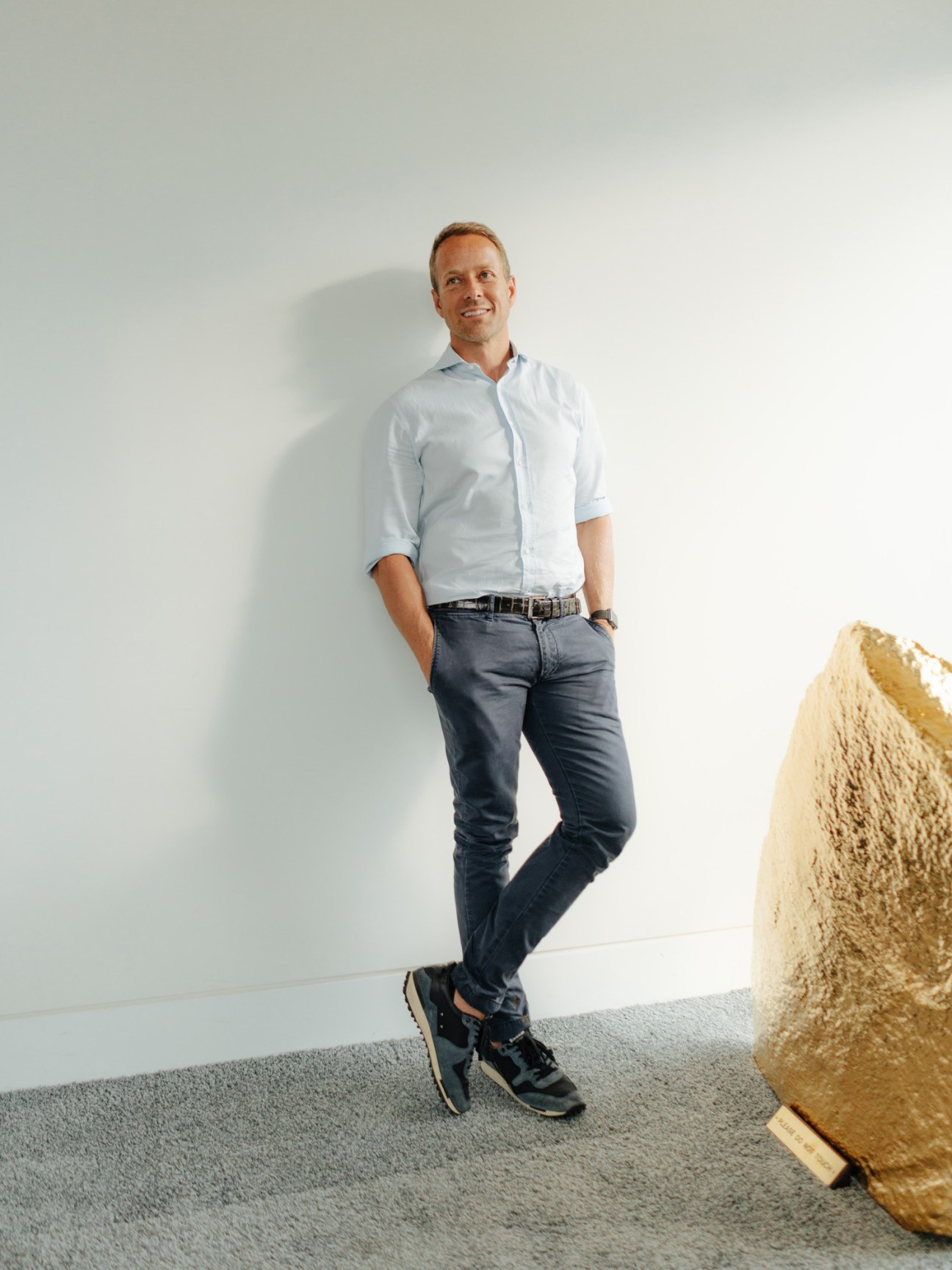 Man in light blue shirt and dark jeans standing against white wall next to gold sculpture