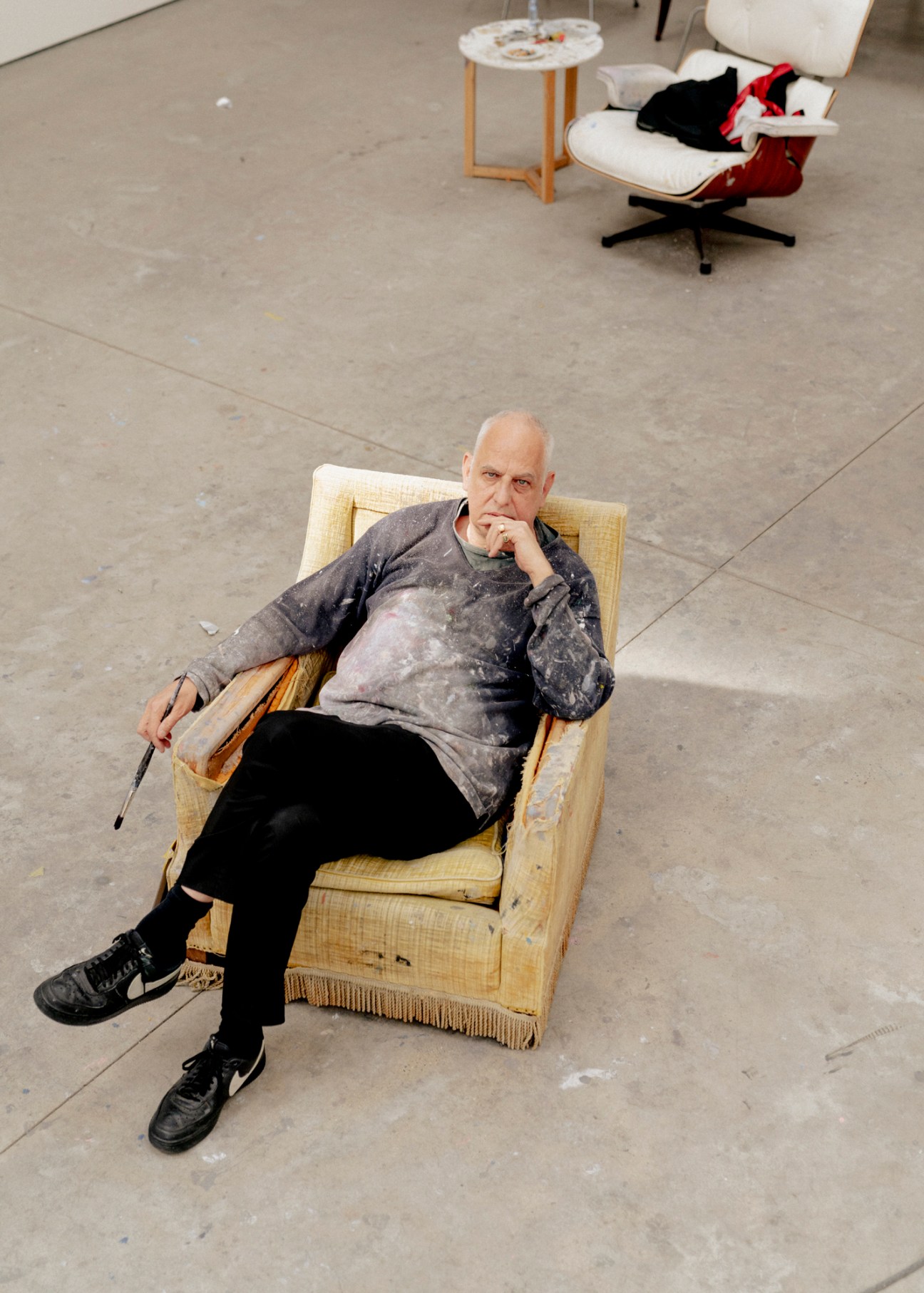 Luc Tuymans sitting in a yellow armchair in his studio with paint-stained clothing and paintbrush in hand