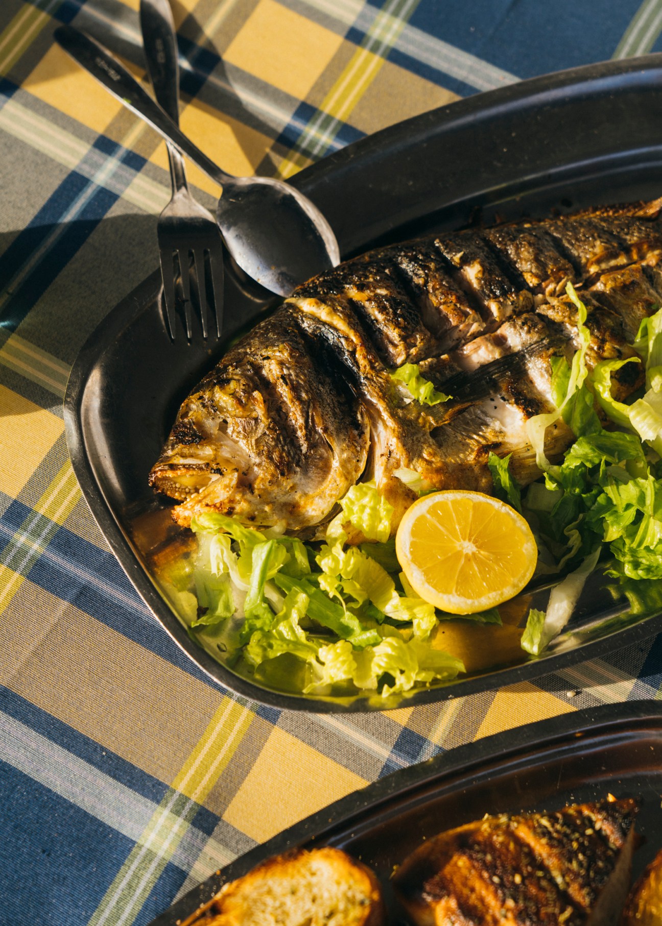 Grilled fish served in a black pan with lettuce garnish and lemon slice, with cutlery on plaid tablecloth