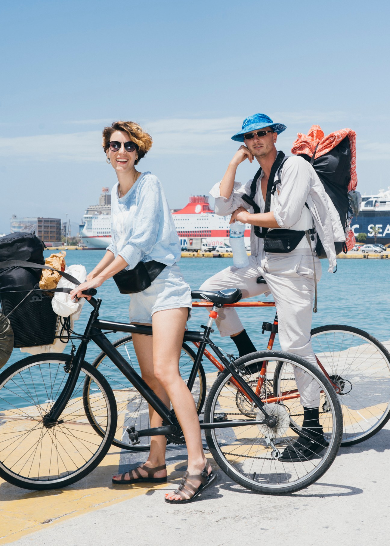 Two people with bicycles and backpacks posing by a waterfront with a ferry in the background