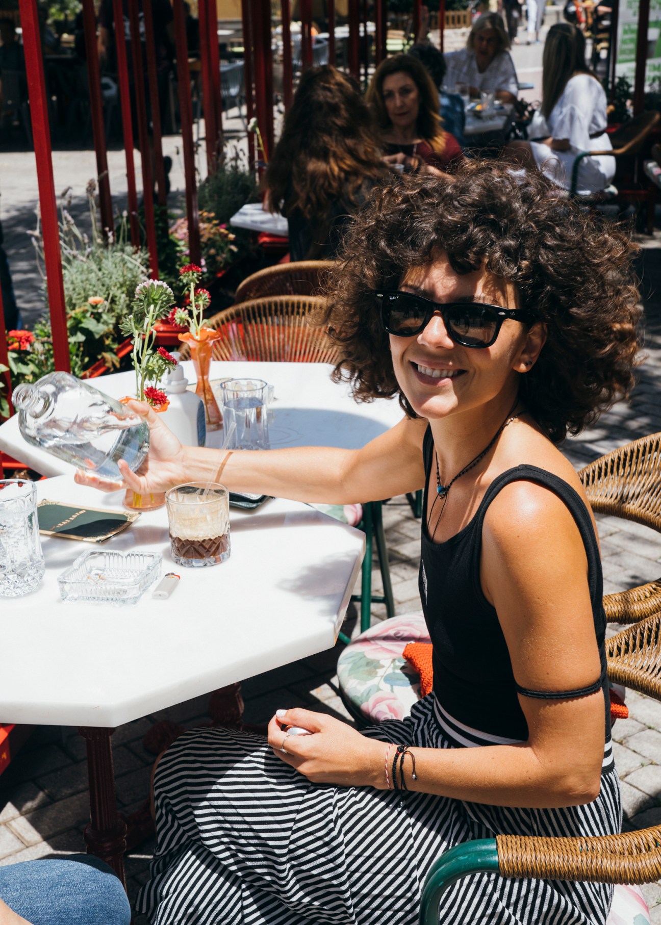 Woman with curly hair and sunglasses smiling while pouring drink at outdoor cafe table