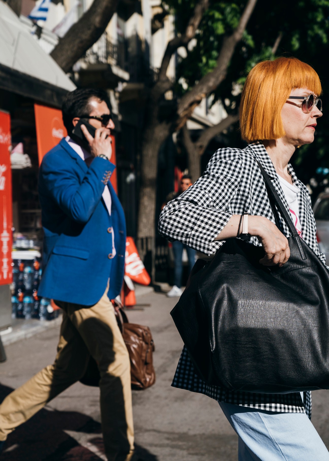 Woman with red hair and sunglasses walks past a man in blue blazer on a tree-lined city street.