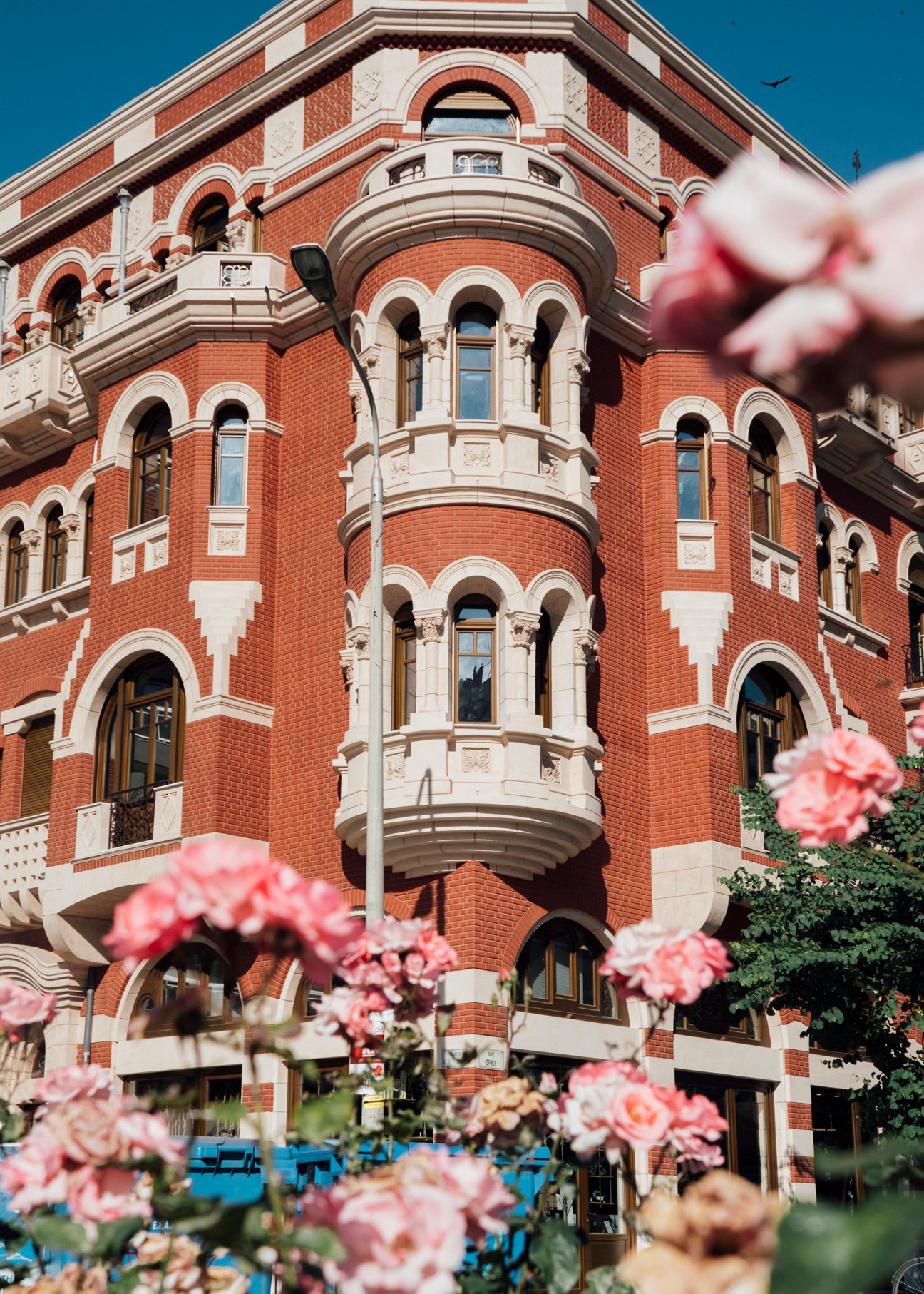 Red brick neobyzantine mansion with ornate white stone archways and pink flowering plants in foreground.