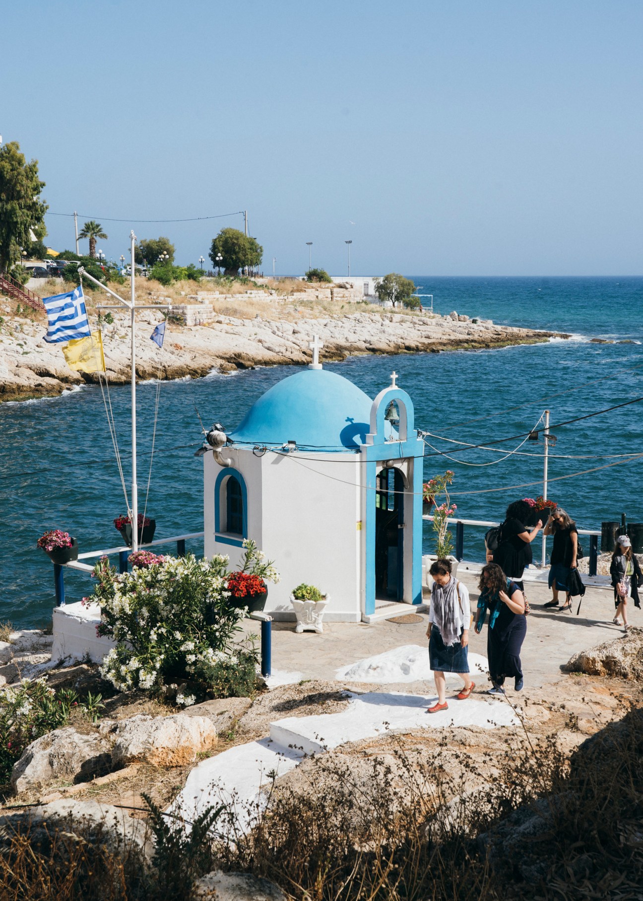White Greek Orthodox church with blue dome and cross sits on rocky coastline overlooking blue sea