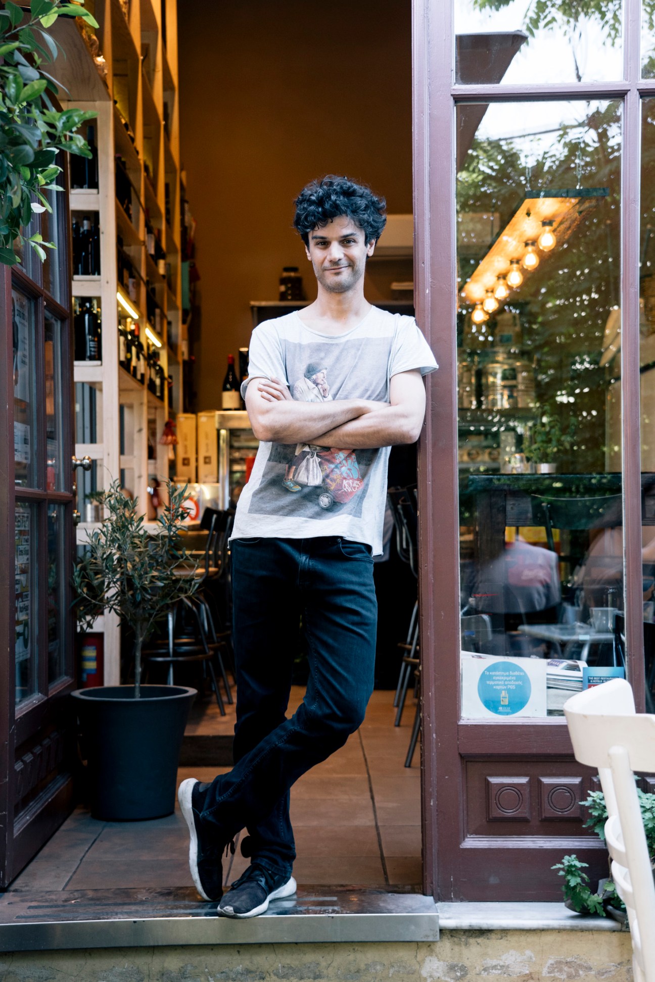 Ilias Agoropoulos stands in the doorway of his restaurant with arms crossed, wearing a light gray t-shirt and dark jeans.