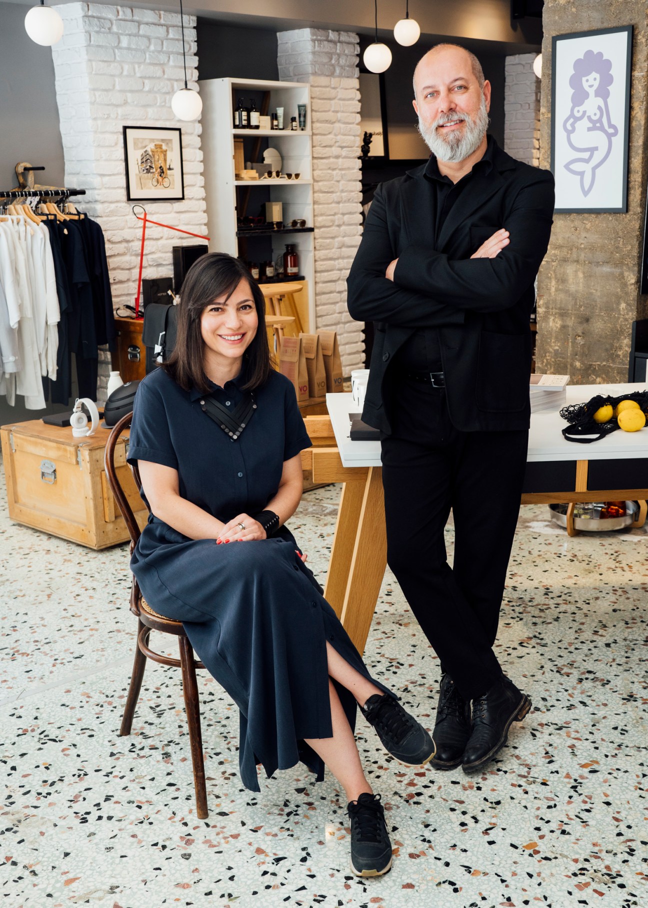 Two people posing in a minimalist boutique shop with white brick walls and shelving displaying products.
