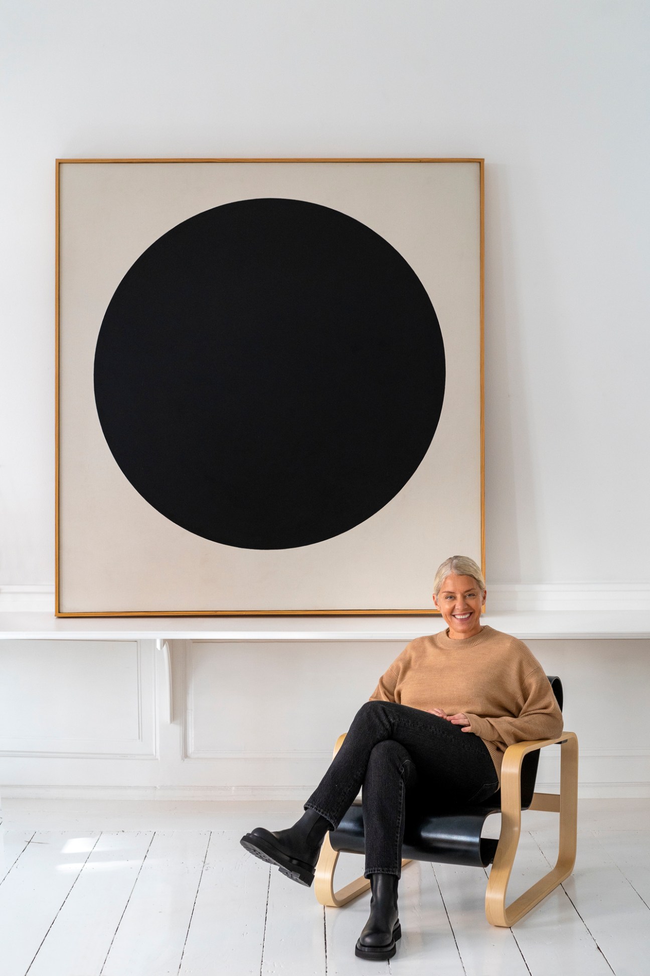 Woman sitting in a wooden chair in front of a large framed artwork with a black circle on cream background