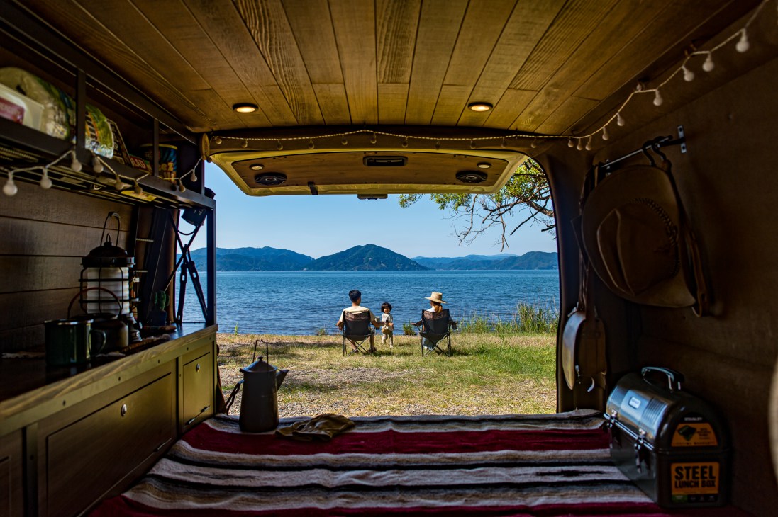View from inside a camper van looking out at two people sitting in chairs by a lake with mountains in the background