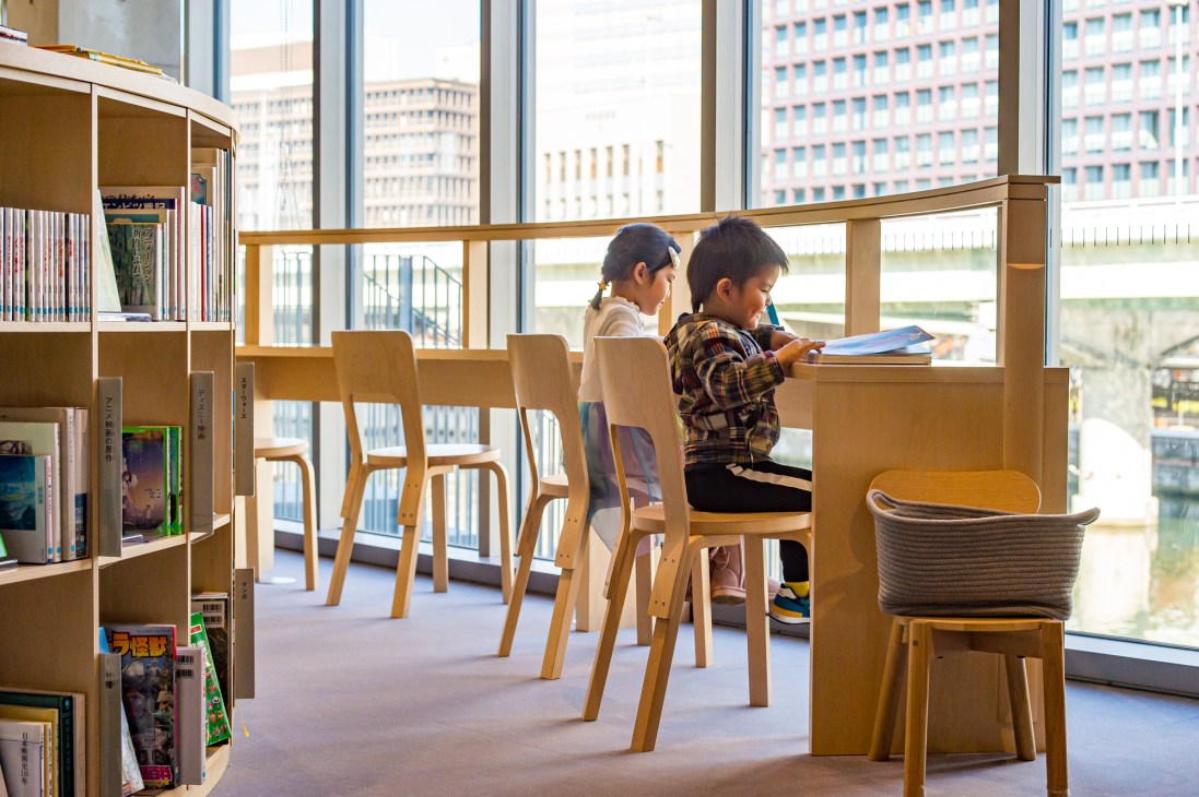 Two children reading at wooden table with Artek N65 chairs in modern library with floor-to-ceiling bookshelves