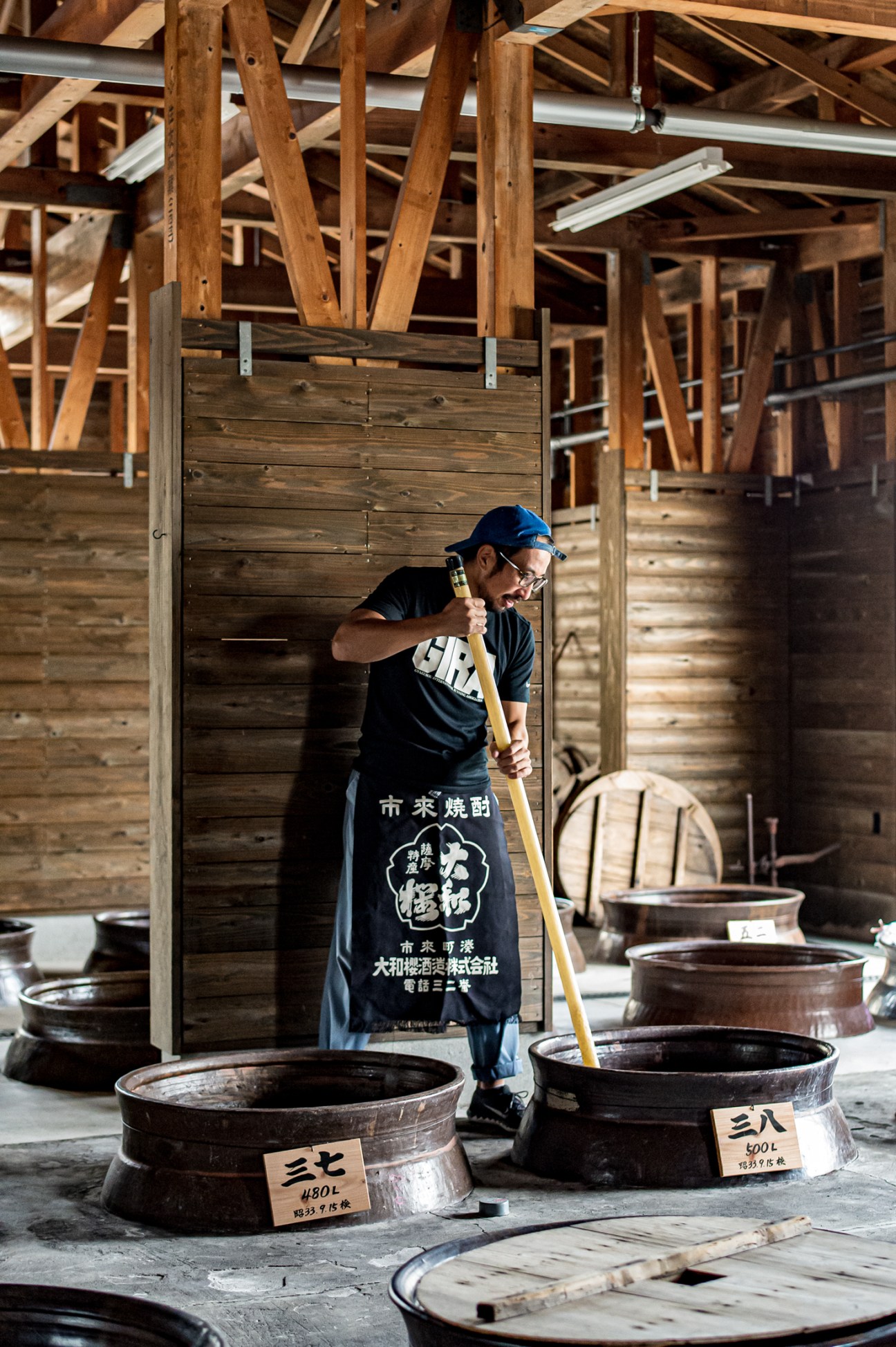 Man in apron stirring large wooden vats with long paddle in traditional wooden distillery building