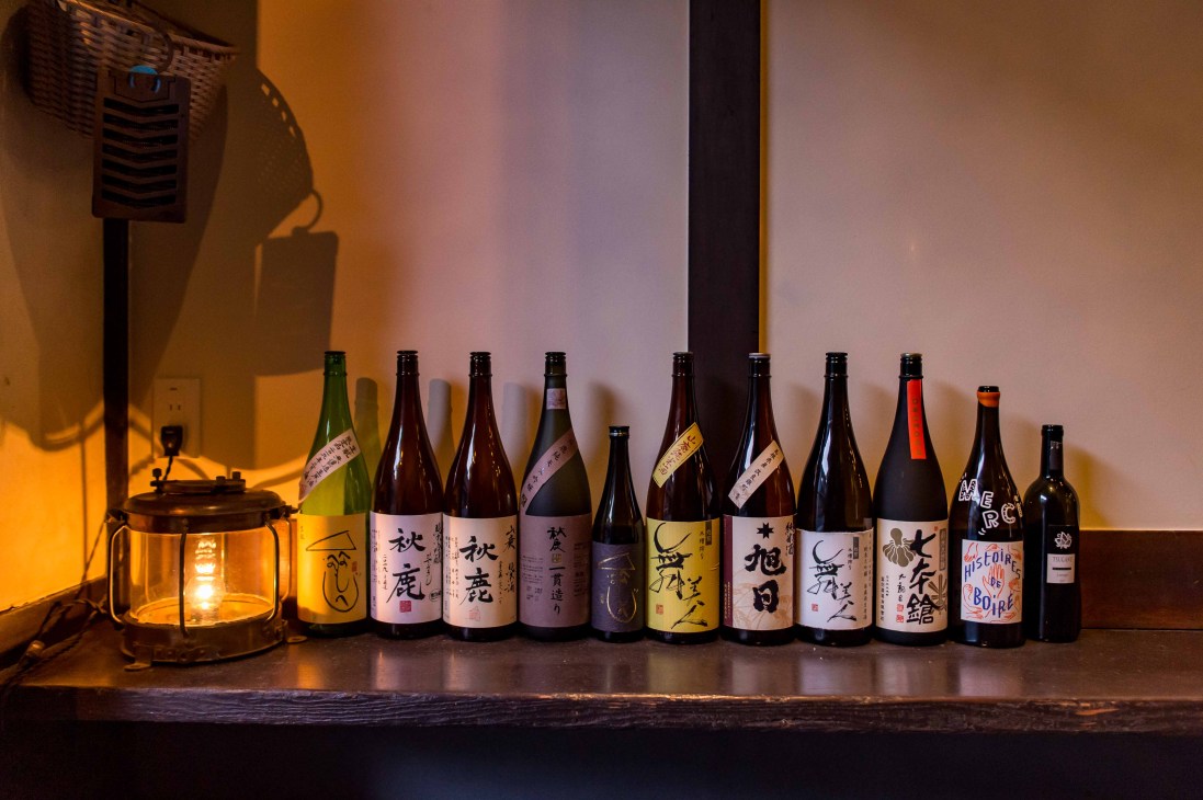 Row of sake bottles with Japanese labels lined up on wooden shelf with warm ambient lighting