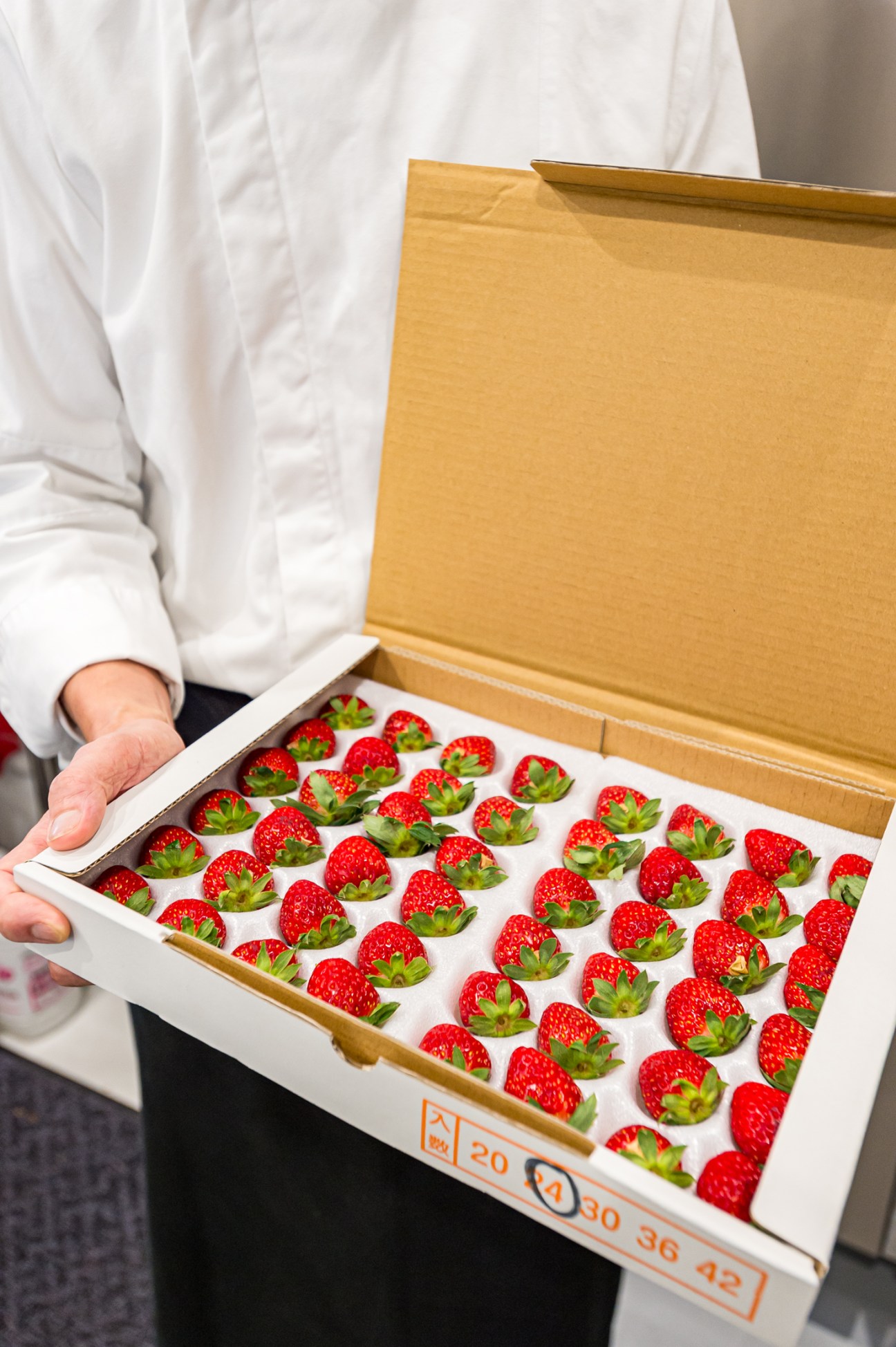 Person in white chef's coat holding open a gold box displaying rows of fresh red strawberries with green stems on white paper.