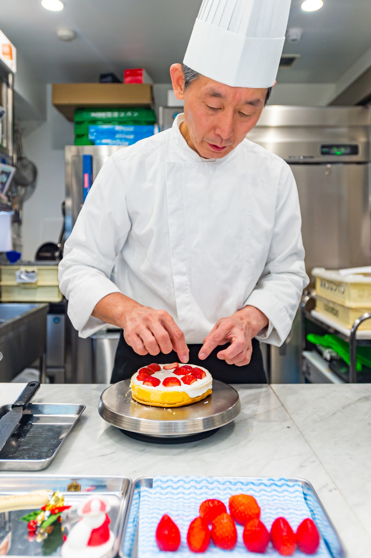 Chef in white uniform and hat decorating a strawberry shortcake with fresh red strawberries in a professional kitchen.