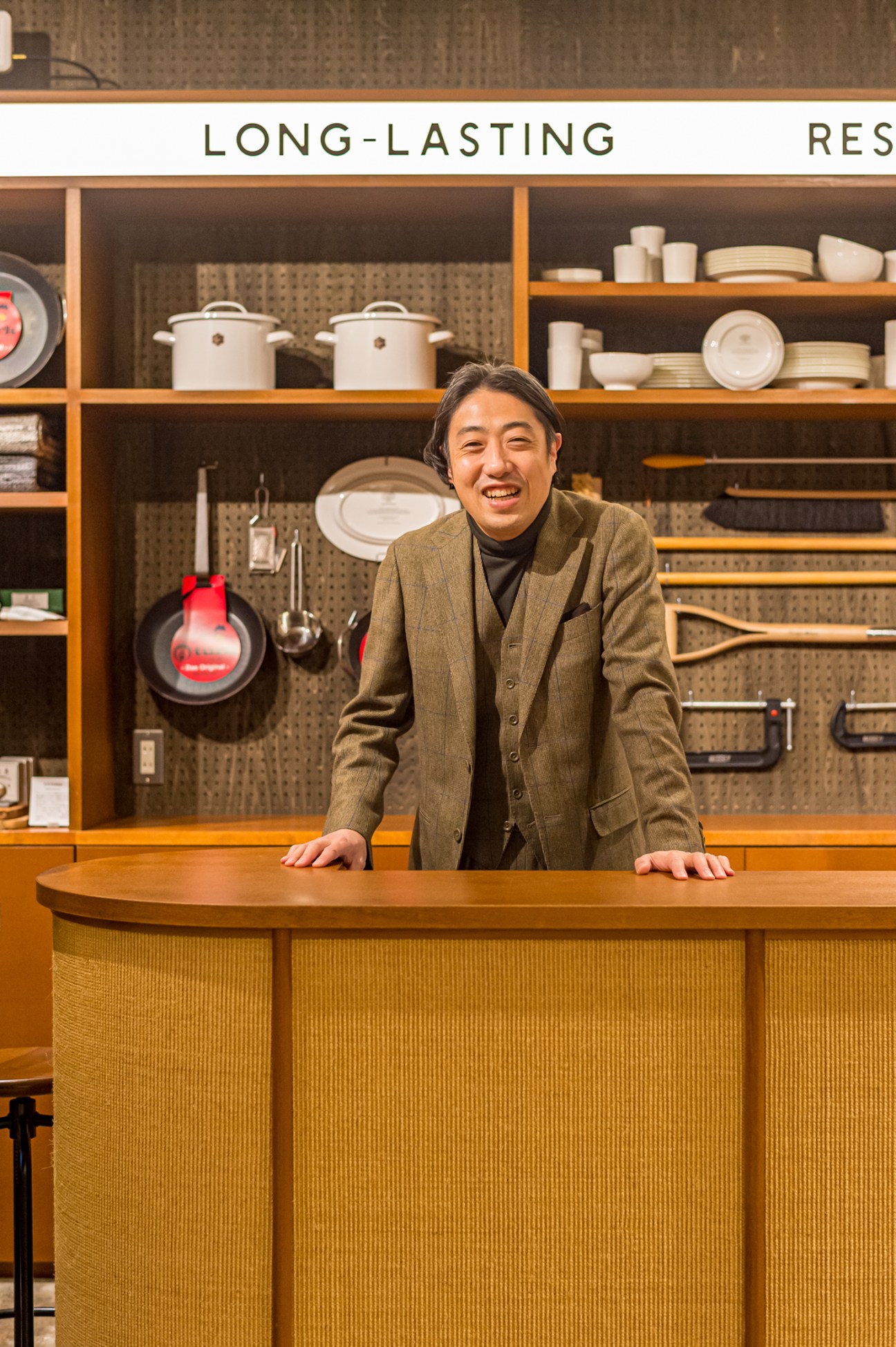 Man in brown suit standing behind curved counter in kitchen store with shelves of dishes and cookware