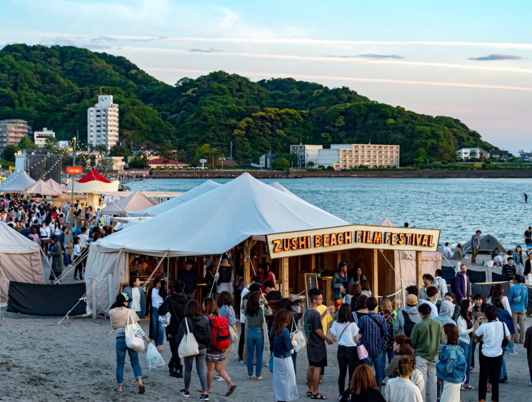 Crowds gather at the Zushi Beach Film Festival at dusk, with tents along the waterfront and tree-covered hills behind.