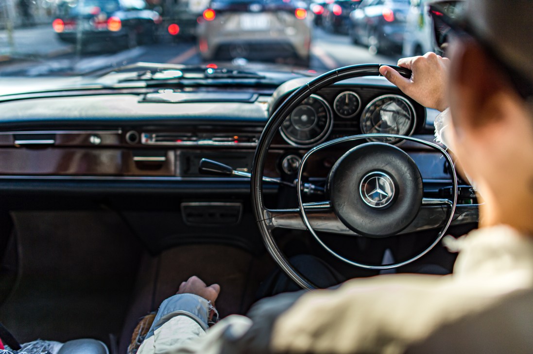Driver's hands on large steering wheel of 1970 Mercedes-Benz W108 with dashboard and traffic visible through windscreen