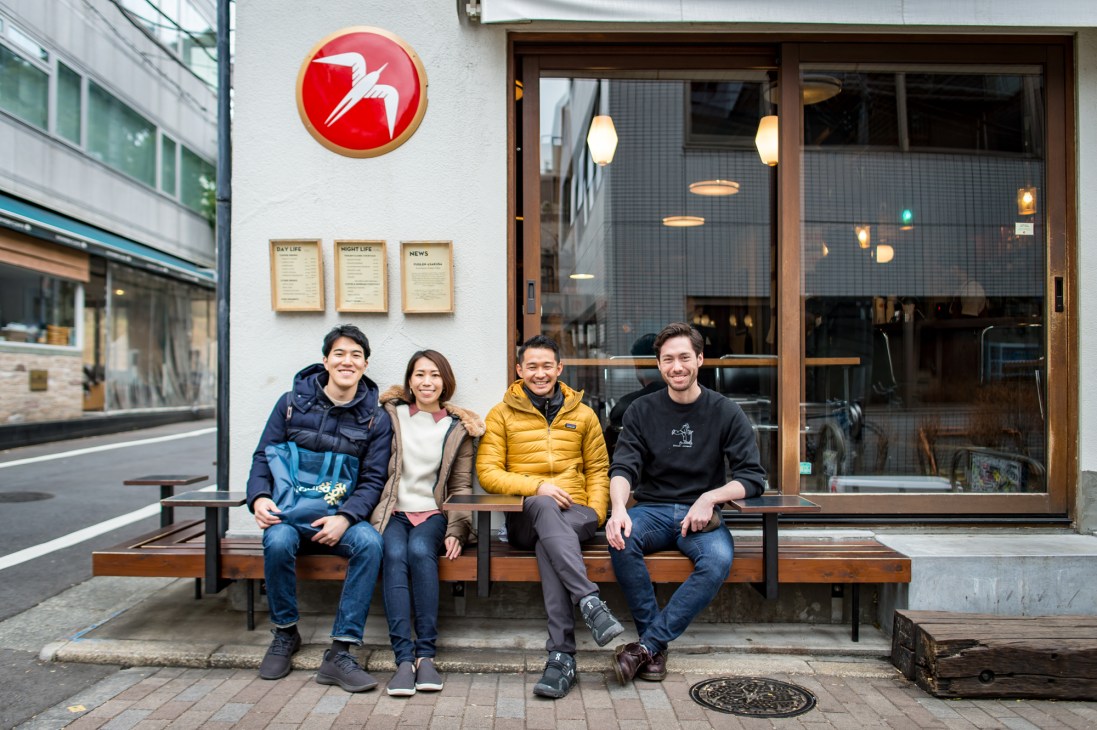 Four people sitting on a wooden bench outside a modern cafe storefront with large windows
