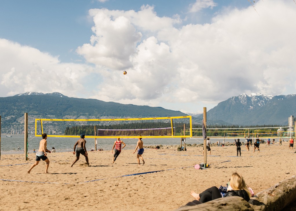 Beach volleyball game with players and spectators on sand court with mountains and water in background