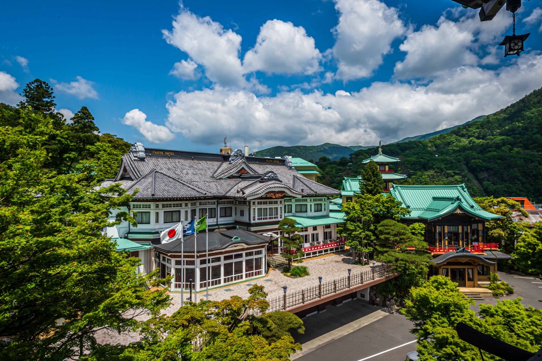 Aerial view of Uniqlo City headquarters with traditional Japanese architecture, surrounded by lush green mountains and blue sky with white clouds.