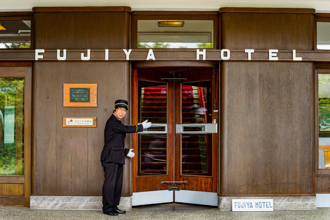 Hotel doorman in black uniform and cap opening ornate wooden doors at Fujiya Hotel entrance