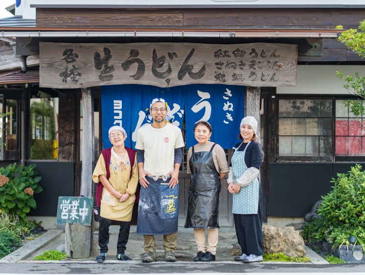 Four staff members smile outside a traditional Japanese udon restaurant with wooden signage and noren curtains.