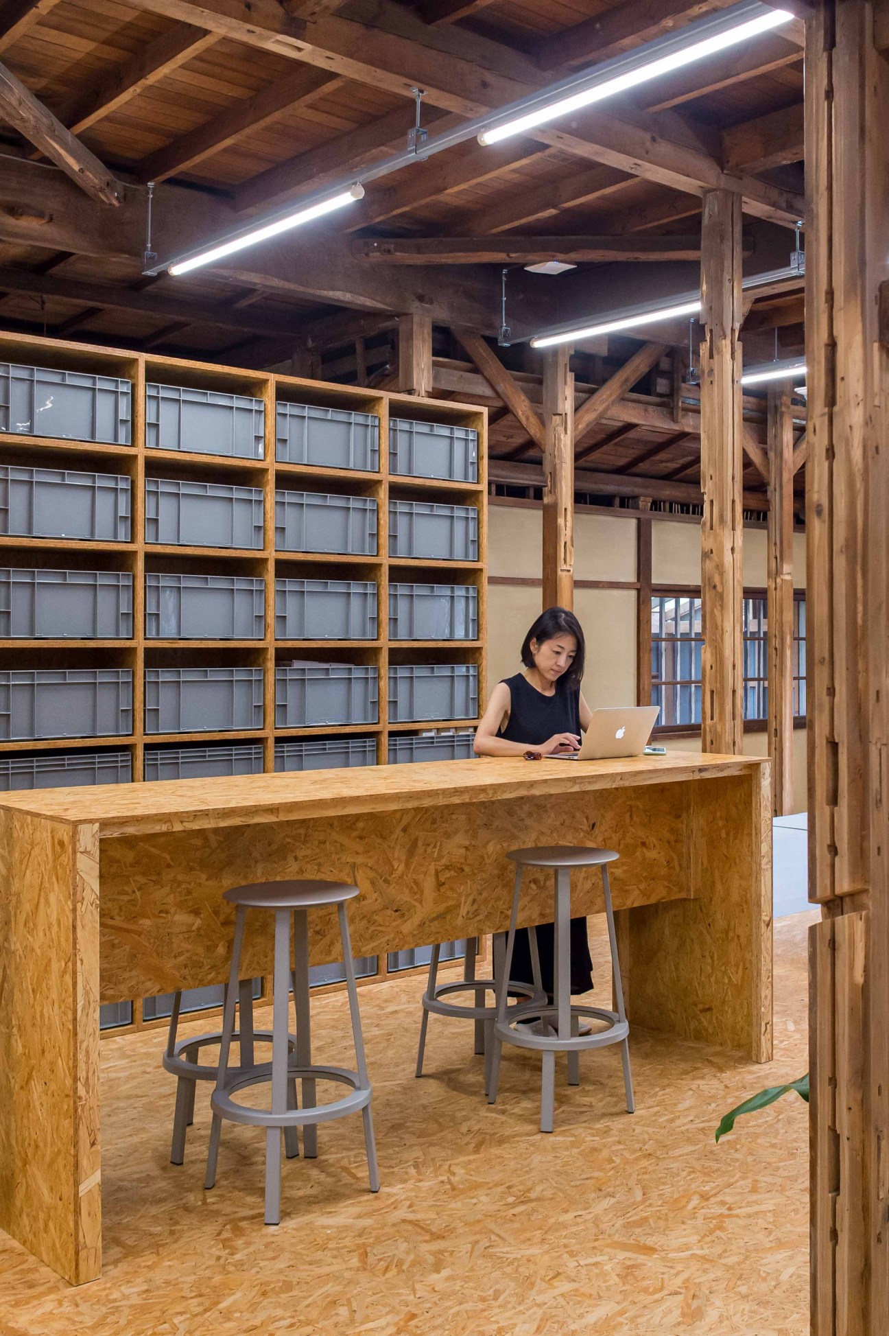 Woman working at a wooden desk with a laptop in a minimalist office space with exposed timber beams and linear ceiling lights.