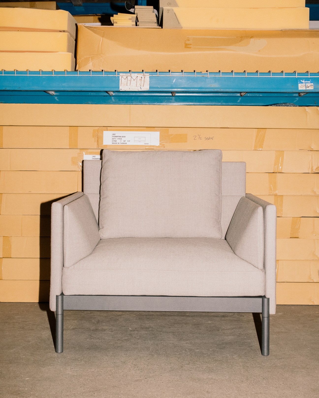 Light grey upholstered armchair with metal legs displayed in a warehouse with cardboard boxes stacked behind.