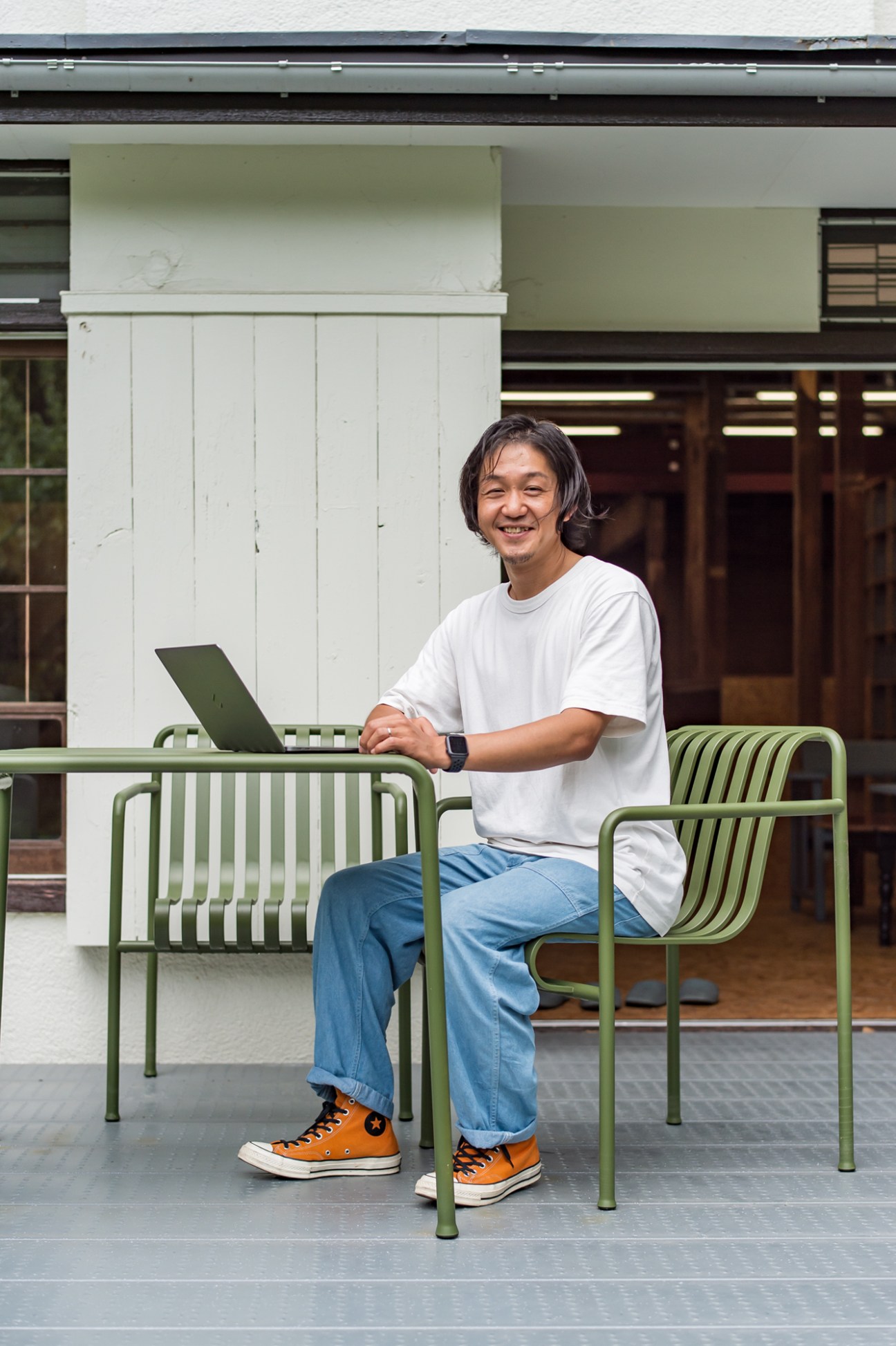 Man in white t-shirt and jeans sitting on green metal chair working on laptop outdoors.