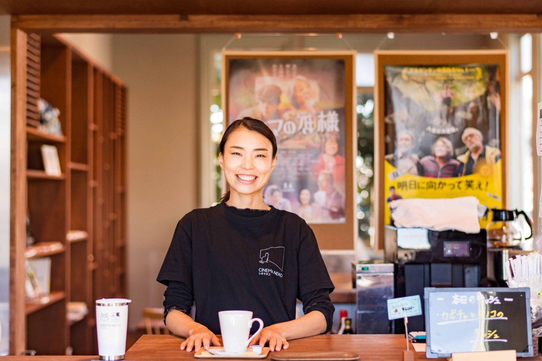Smiling woman in black Cinema Neko shirt standing behind wooden counter at cinema cafe