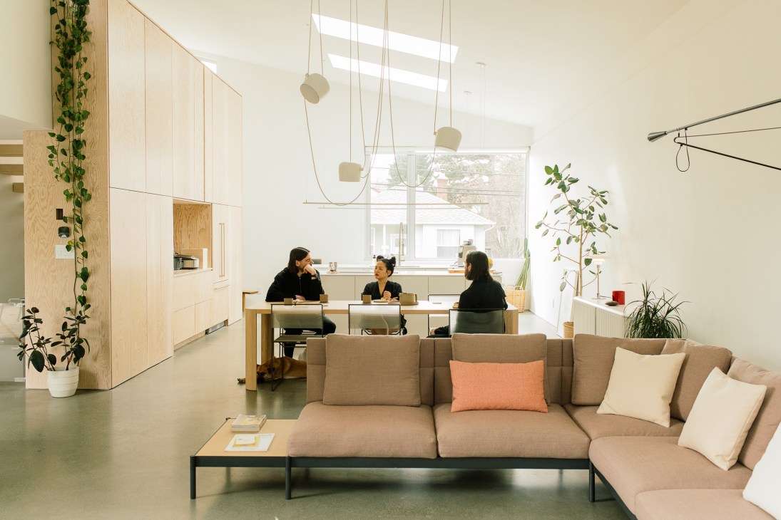 Modern office interior with beige sofa, suspended pendant lights, and three people seated at a wooden desk by a window.