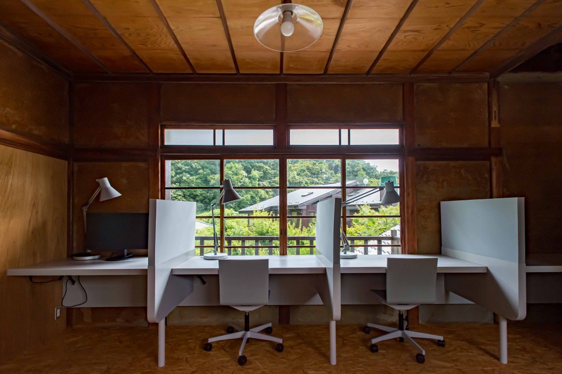 Two white desks with grey office chairs and privacy screens in a wood-beamed office with large windows overlooking a forested landscape.