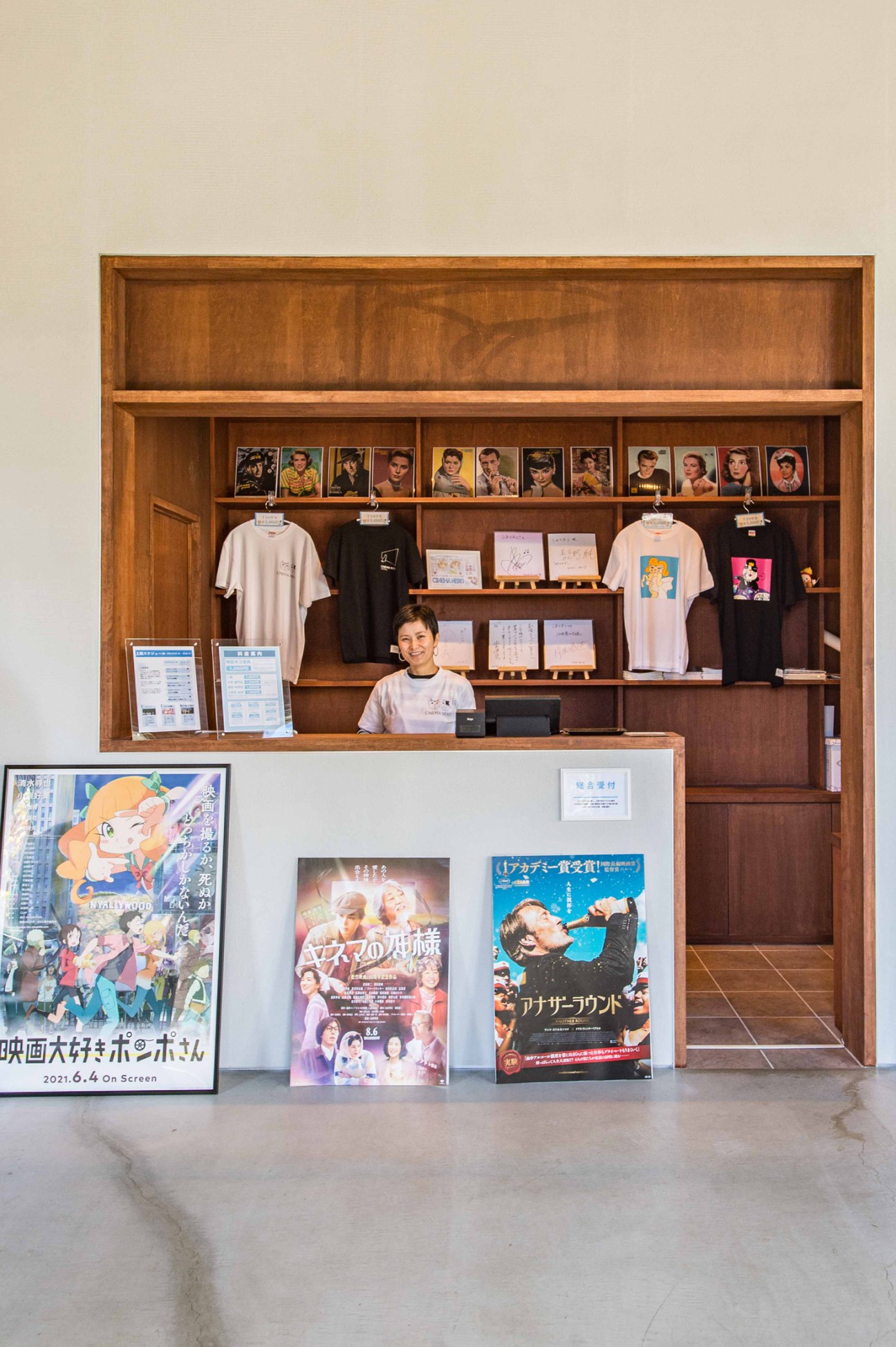 Cinema ticket counter with staff member behind desk, movie posters displayed on walls and merchandise visible on shelves