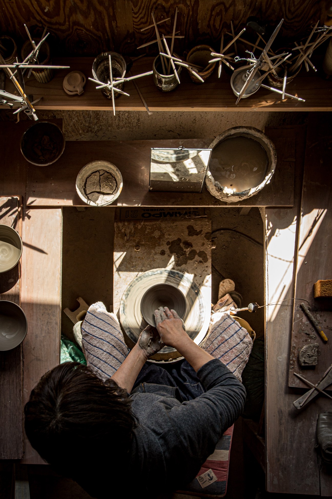 Potter working at a wheel in a rustic studio with ceramic vessels and tools visible.