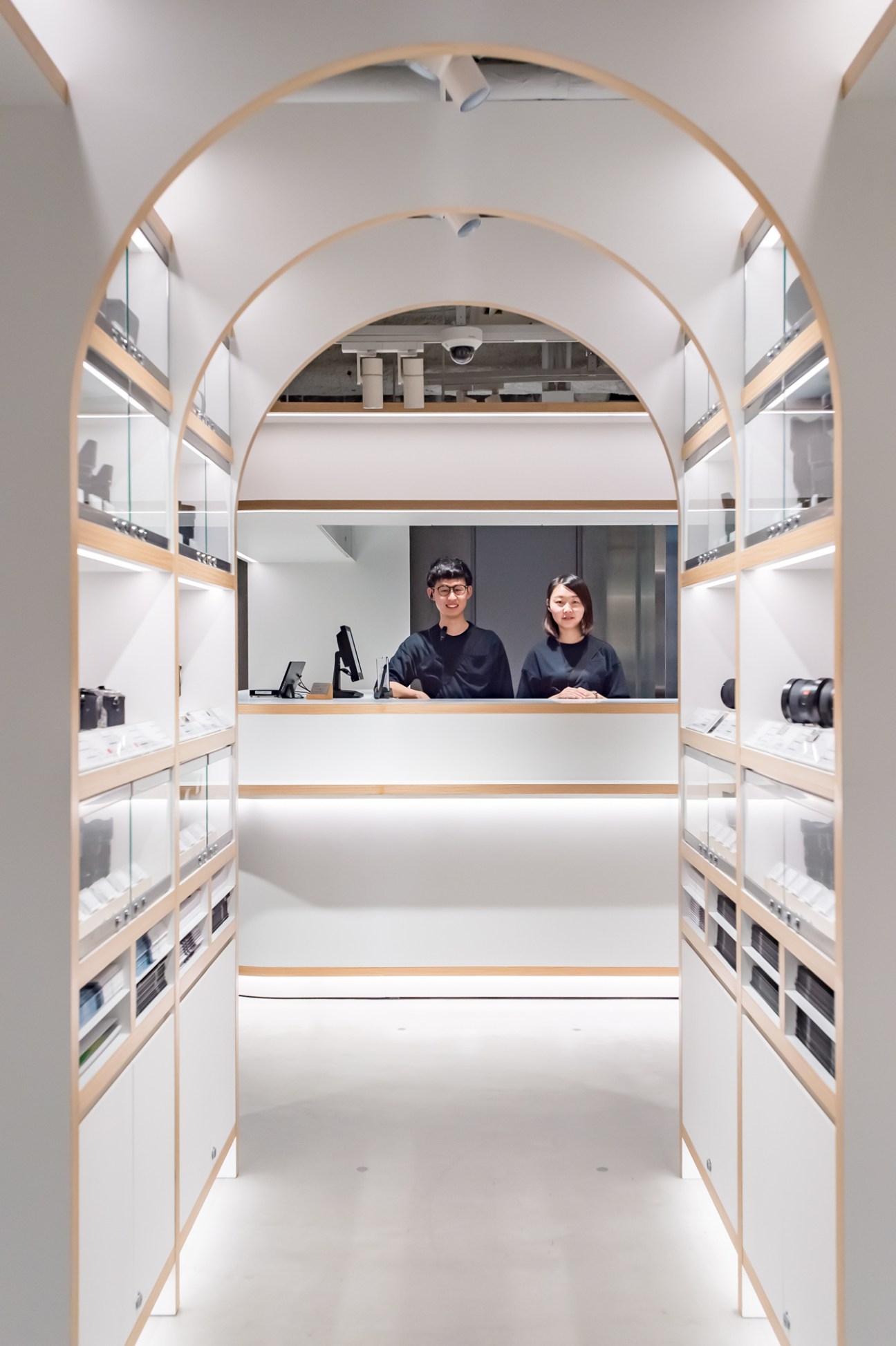 Modern camera store interior with white arched ceiling, wooden shelving displaying cameras, and two staff members at counter