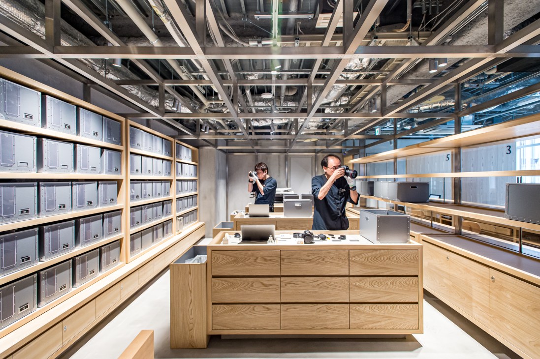 Two technicians working in a glass-walled camera repair studio with exposed ceiling and wooden shelving