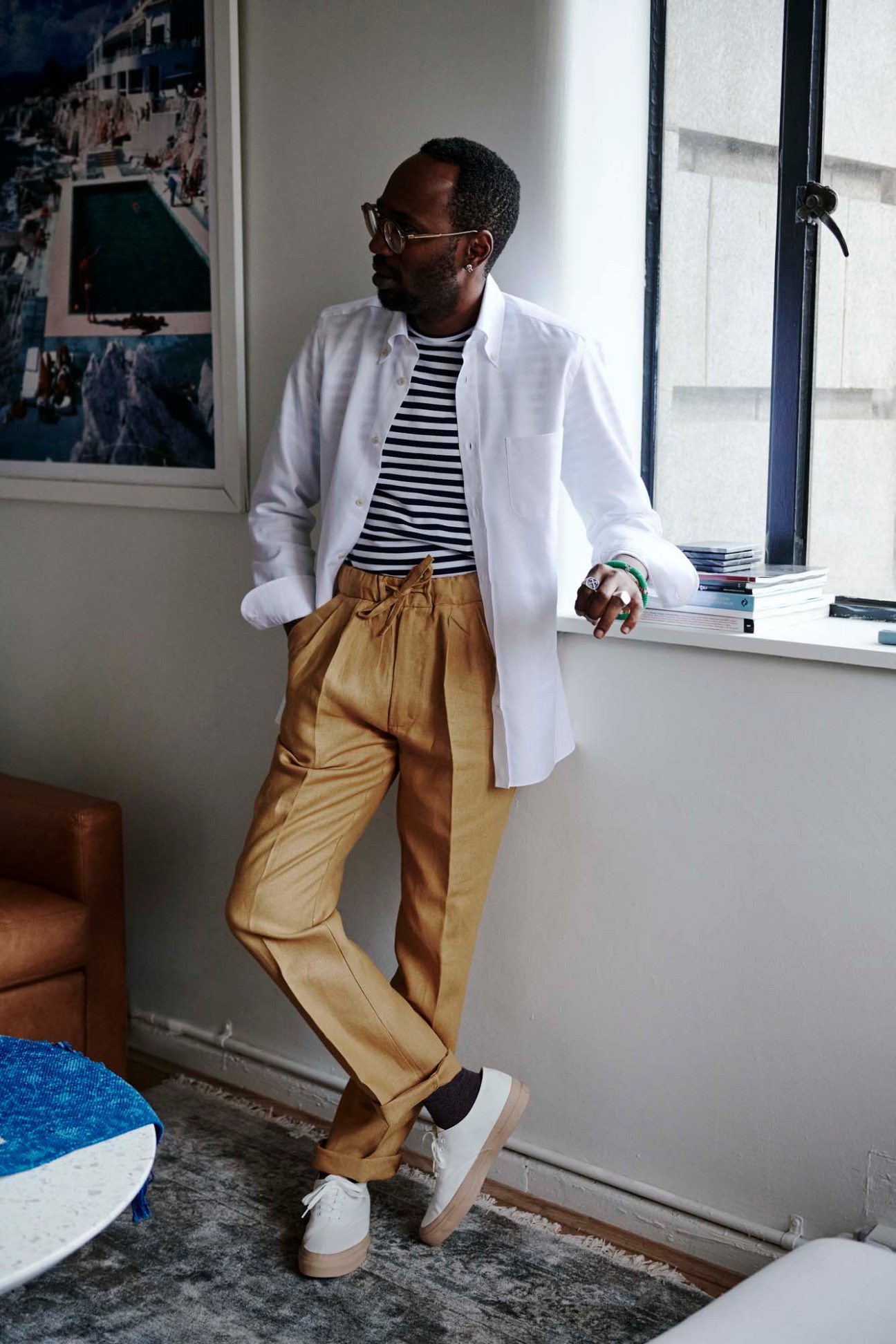 Man wearing white blazer, striped shirt, tan trousers and white sneakers by a window