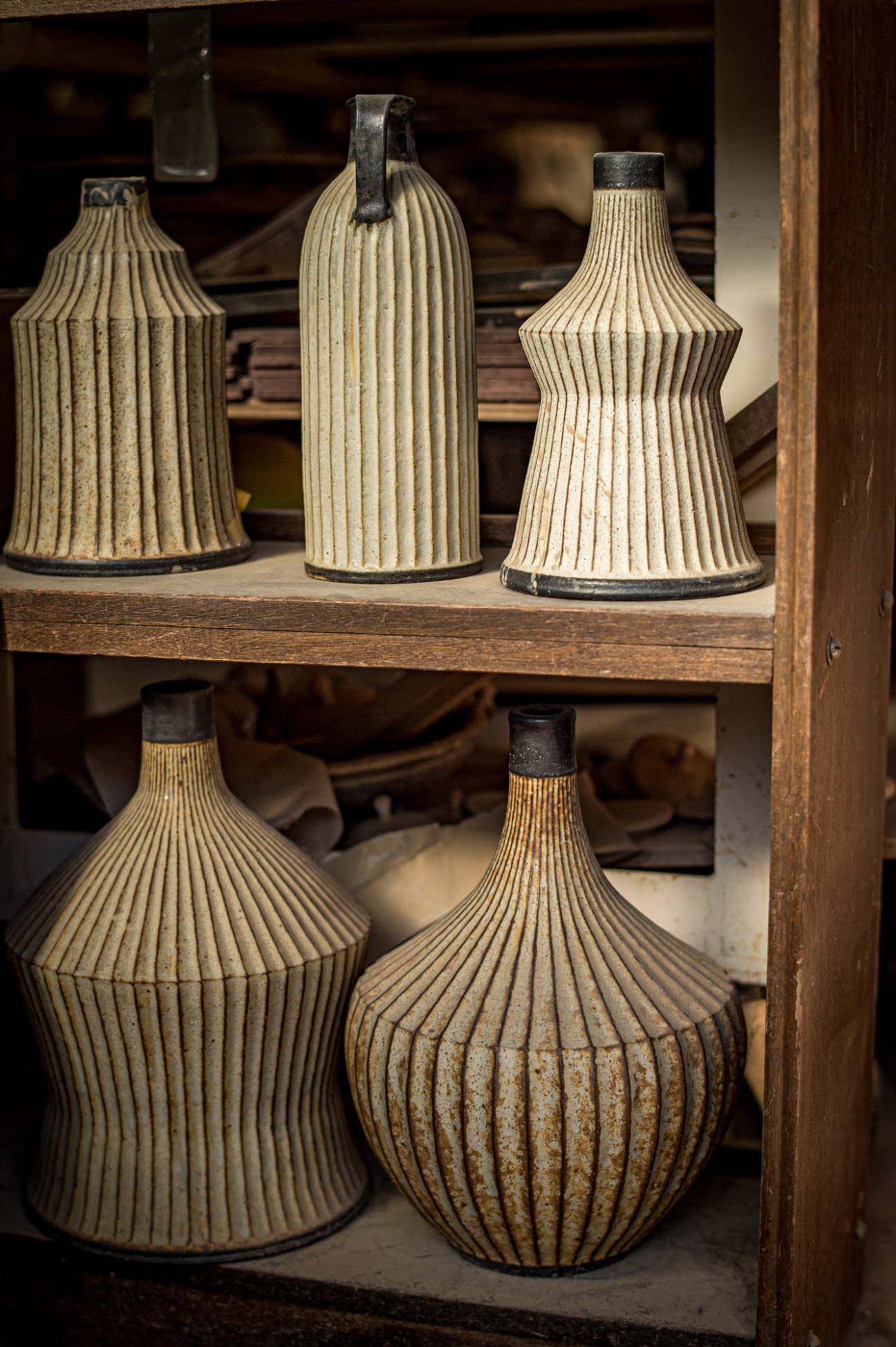 Five ribbed ceramic vessels with dark metal caps displayed on wooden shelves in a studio.