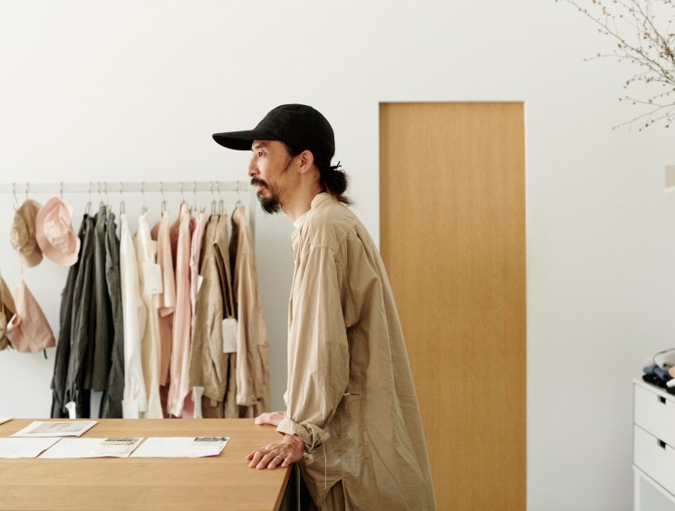 Man in black cap and beige shirt standing in minimalist studio with clothing rack and wooden surfaces