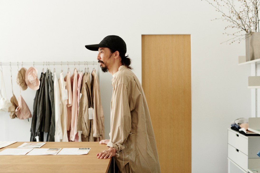 Man in black cap and beige shirt standing in minimalist studio with clothing rack and wooden surfaces