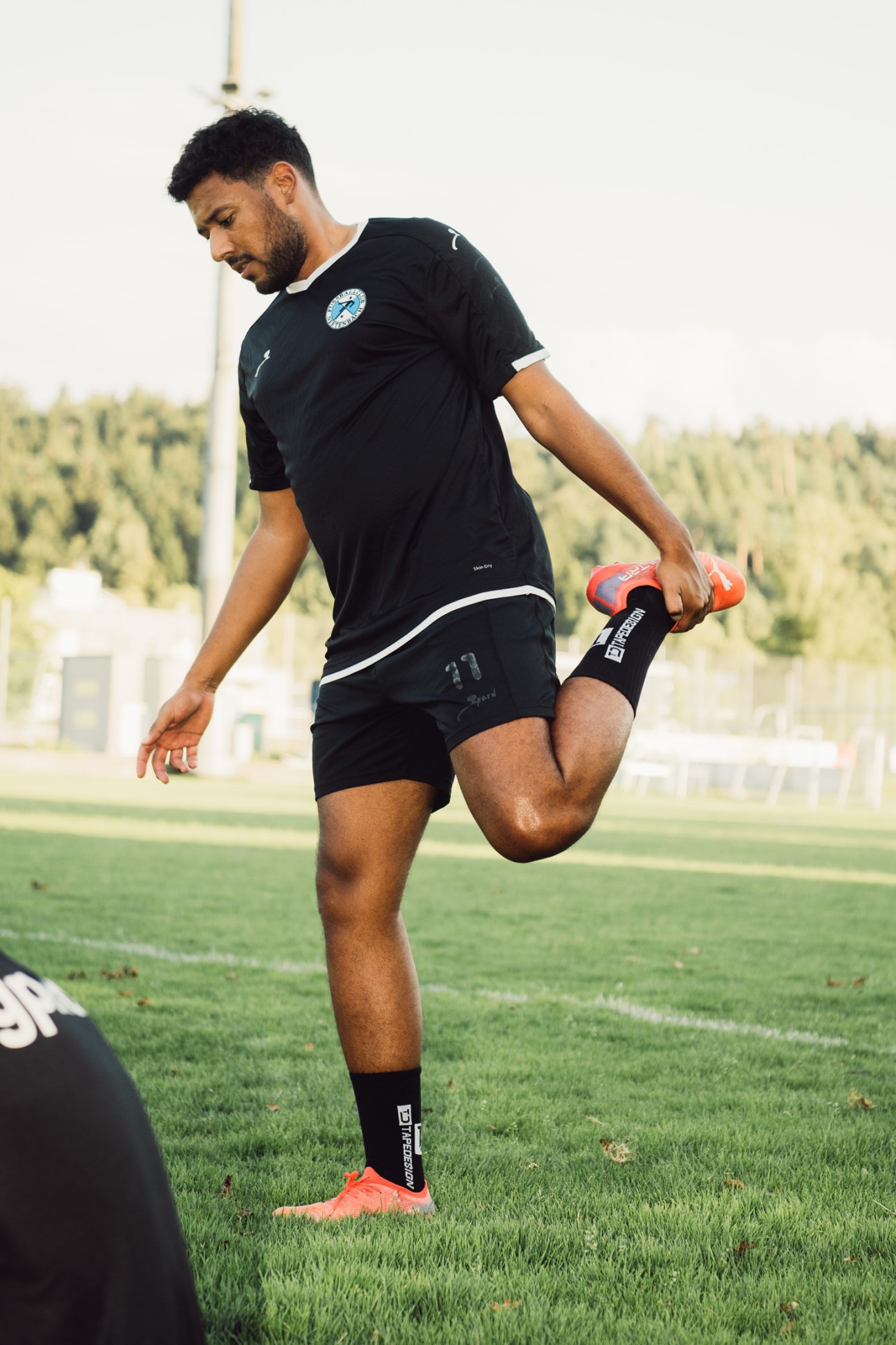 Football player in black uniform stretching his leg on a grass field during training