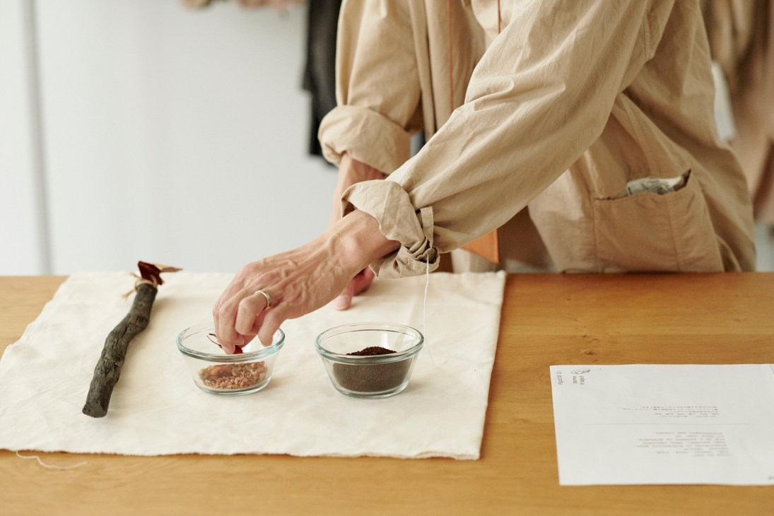 Person in beige linen clothing working with natural dye materials including bowls of coffee grounds and other ingredients