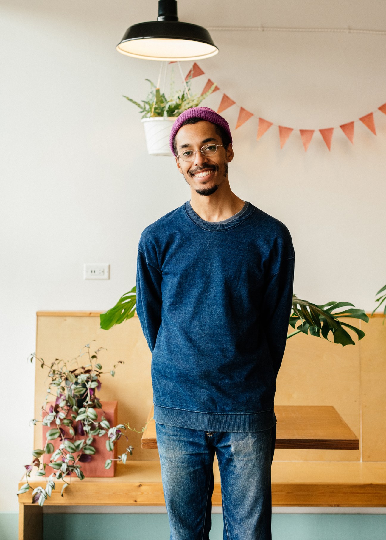 Man in purple beanie and blue sweatshirt smiling in bright studio space with black pendant lamp, potted plants and orange bunting